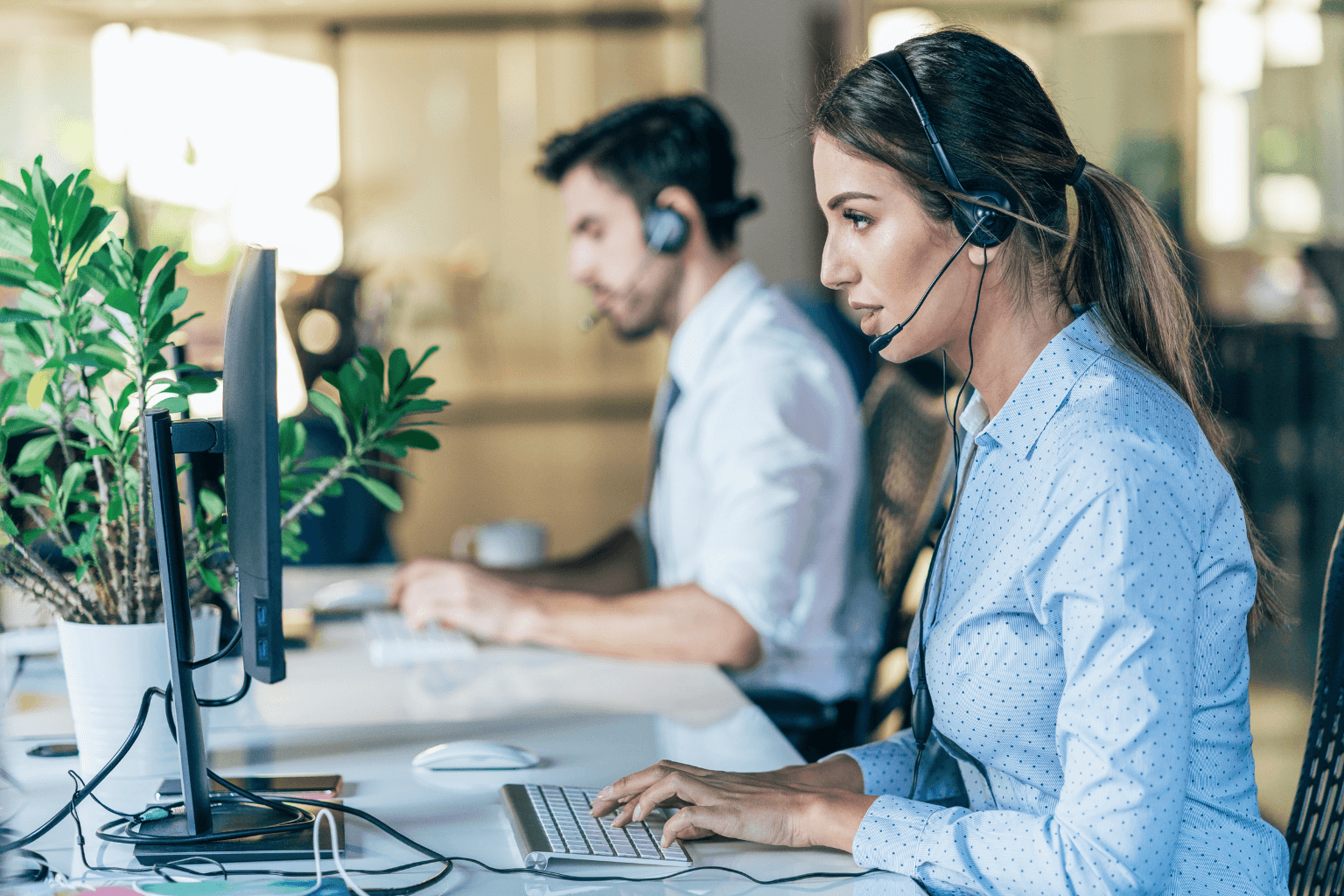 A woman and a man wearing headsets work at their computers in a bright open-plan office.