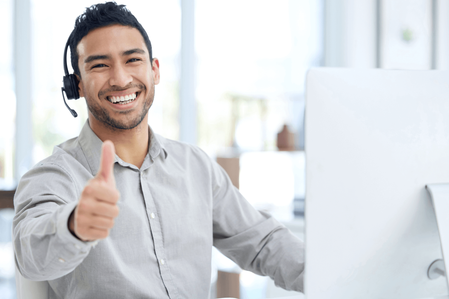 Smiling man wearing a headset gives a thumbs up while seated at a desk with a computer in an office.