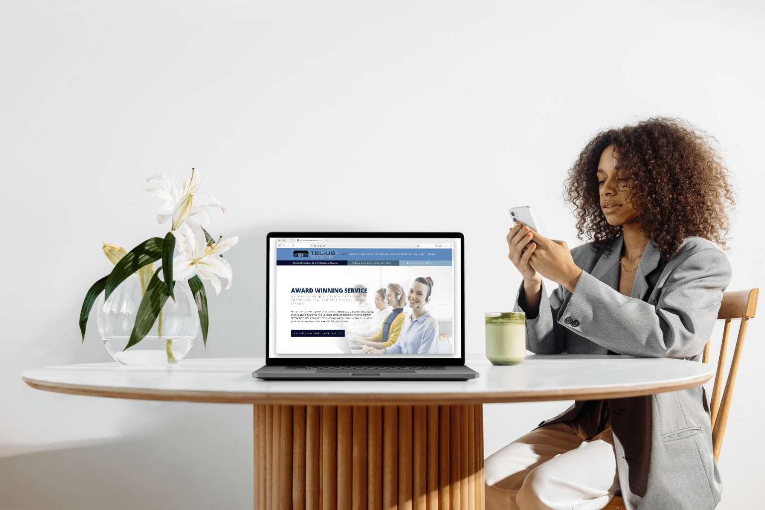 A woman with curly hair wearing a gray blazer sits at a round wooden table, using a smartphone beside a laptop displaying a website, with a vase of white lilies and a candle nearby.