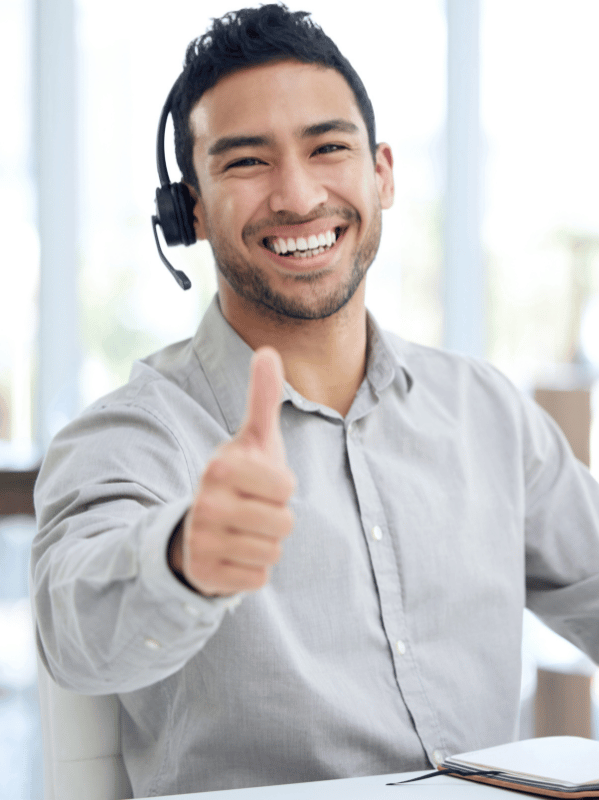 Smiling man wearing a headset gives a thumbs up while seated at a desk in an office.