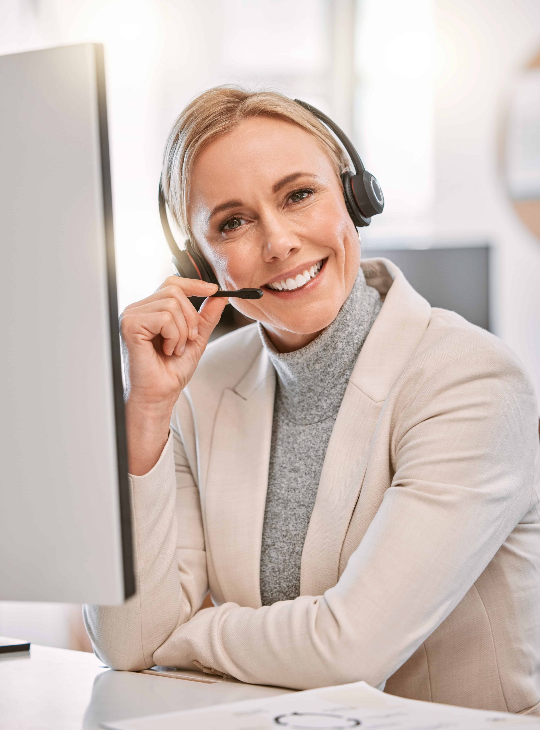 Smiling woman wearing a headset and blazer sits at a desk in an office.