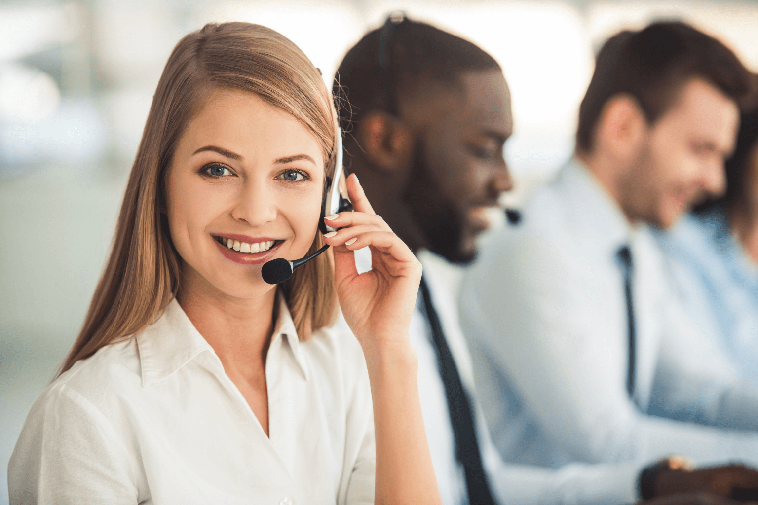 Smiling woman wearing a headset at a customer service desk, with two blurred colleagues in the background.