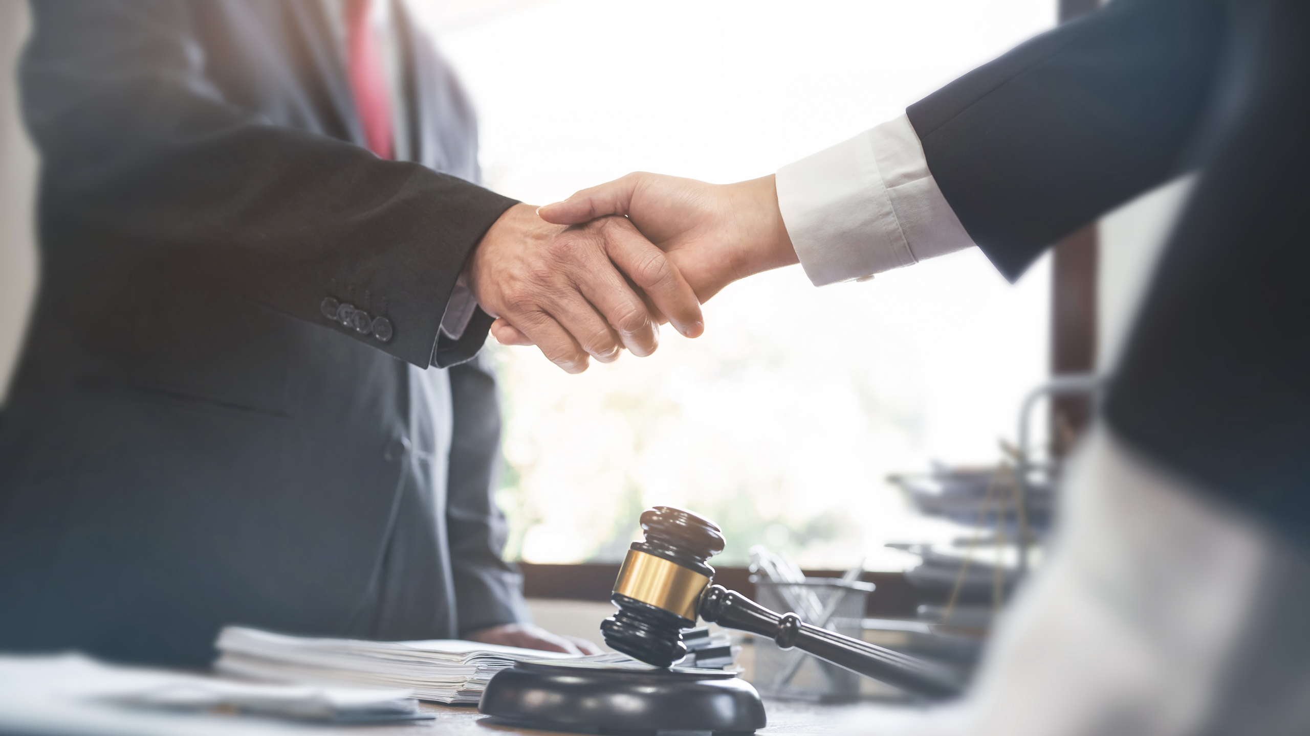 Two professionals in suits shake hands over a desk with a gavel and documents nearby.