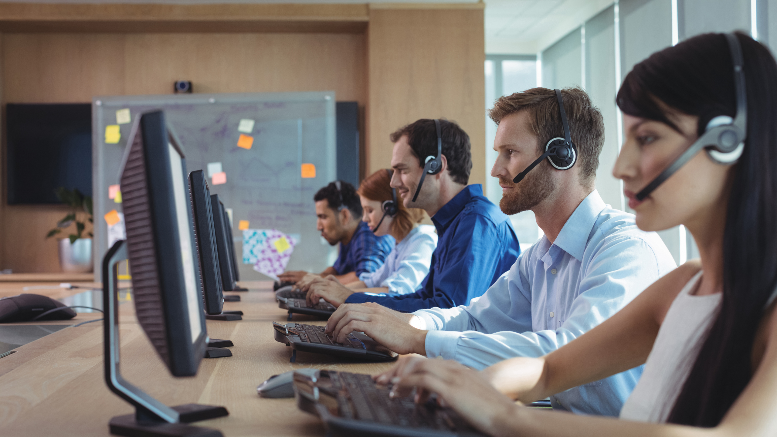 A row of customer service agents wearing headsets sit at computer desks in a modern office.