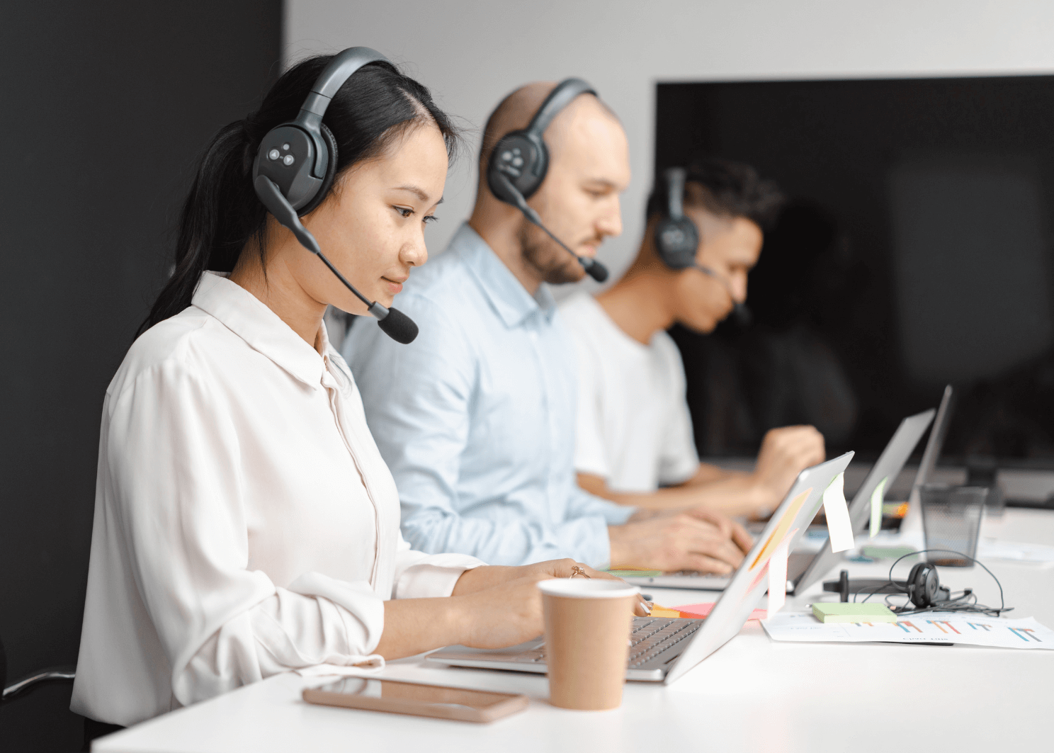 Three call center agents wearing headsets work at laptops in an office.
