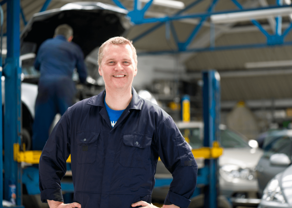 Smiling mechanic in a navy blue coverall stands with hands on hips inside an auto repair shop, with a car on a lift in the background.