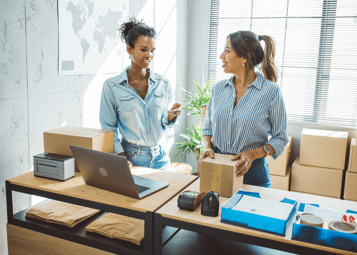 Two coworkers packing boxes at a desk with a laptop and shipping supplies in a bright office.