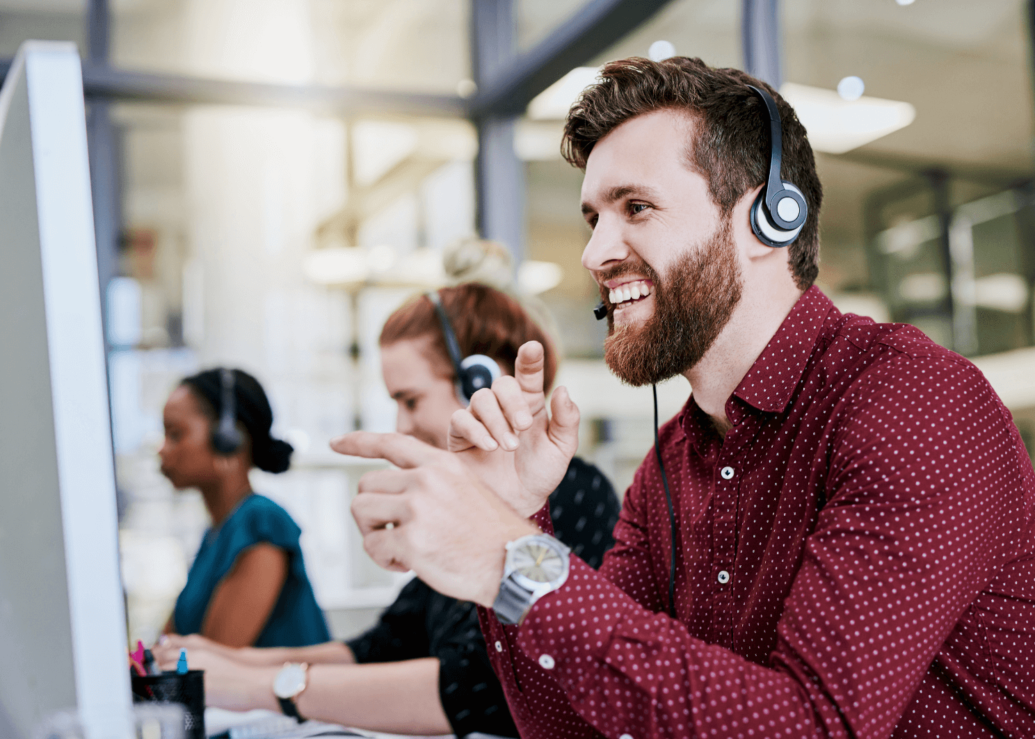 A bearded man wearing a headset gestures and talks at his computer in a busy call center with coworkers in the background.