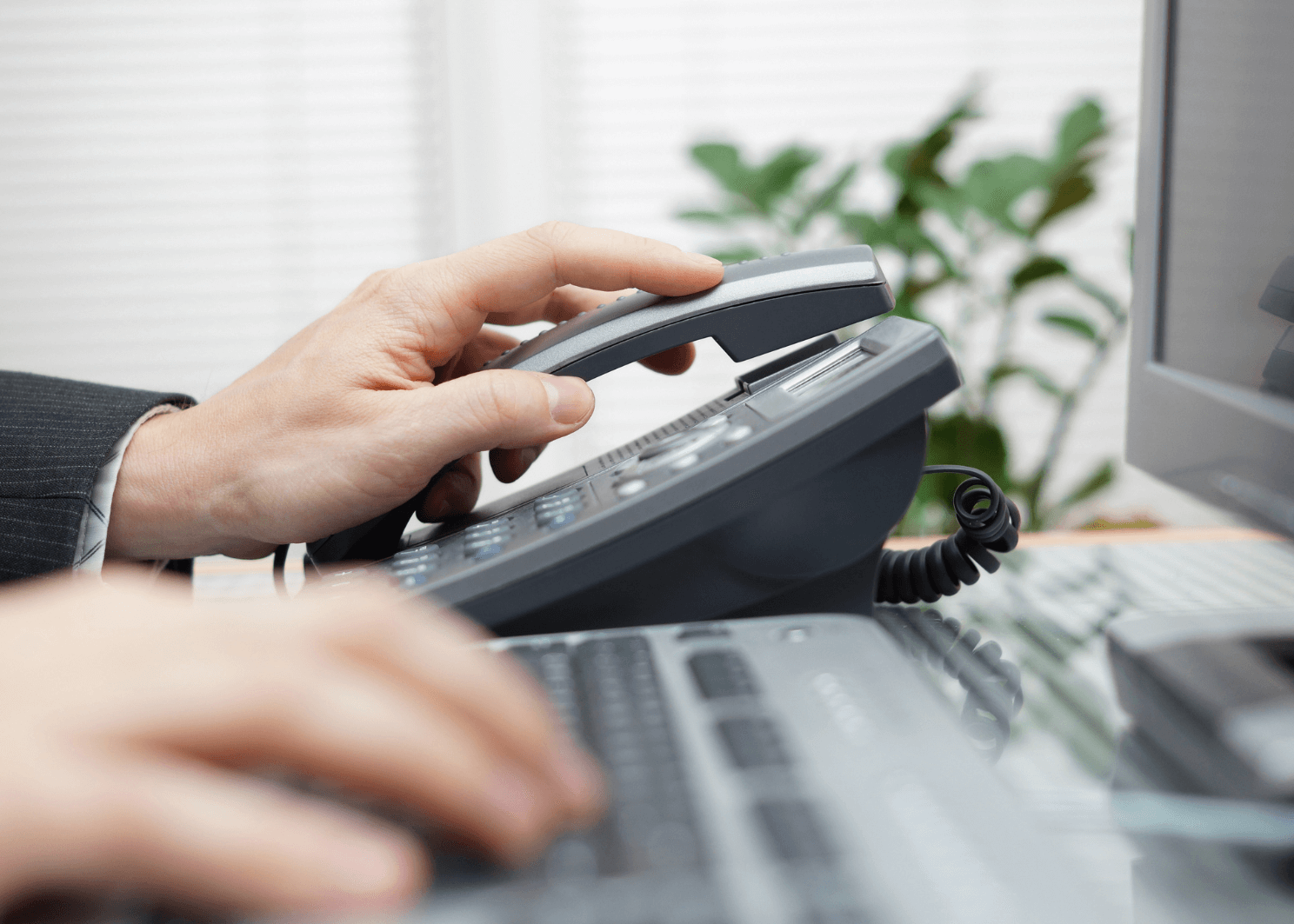 Close-up of a hand pressing buttons on a gray office landline phone beside a computer keyboard.