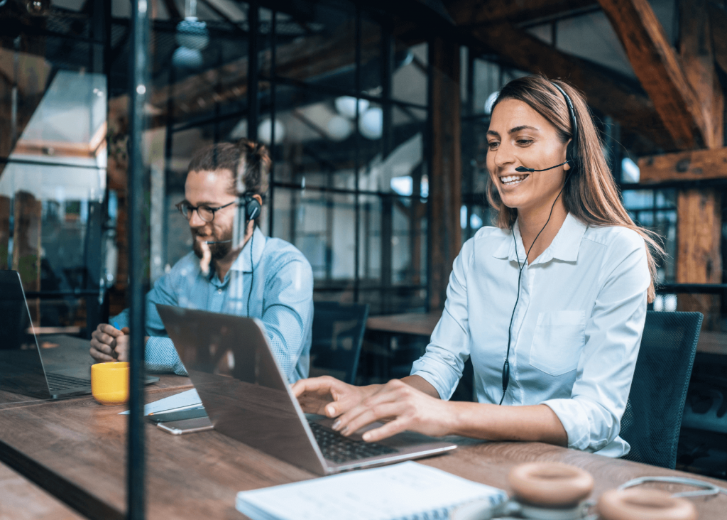 Two colleagues wearing headsets work on laptops at a long wooden table in a bright, modern office.