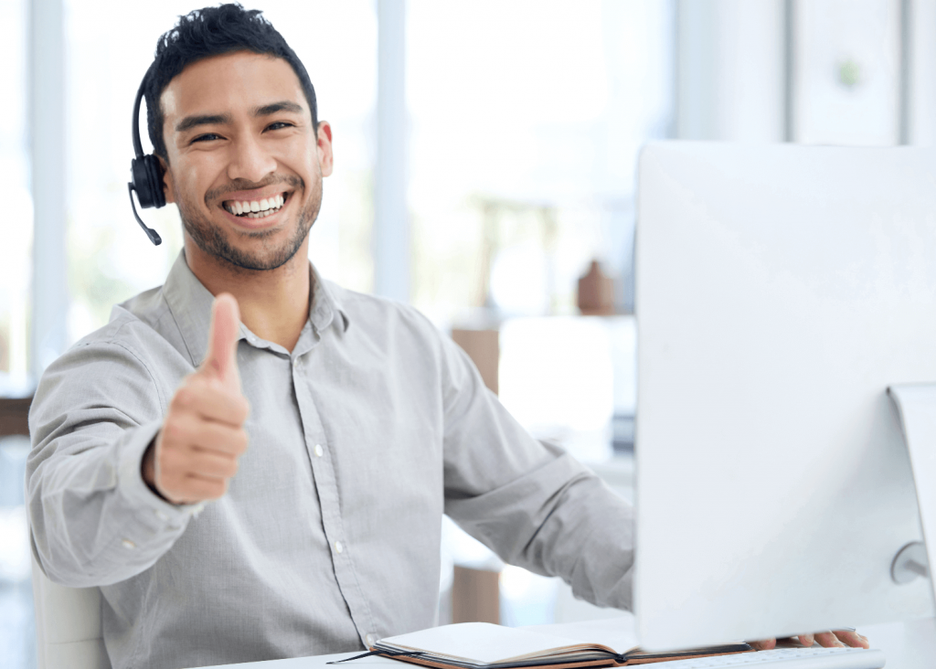 Smiling man wearing a headset sits at a desk with a computer in a bright office, giving a thumbs up.