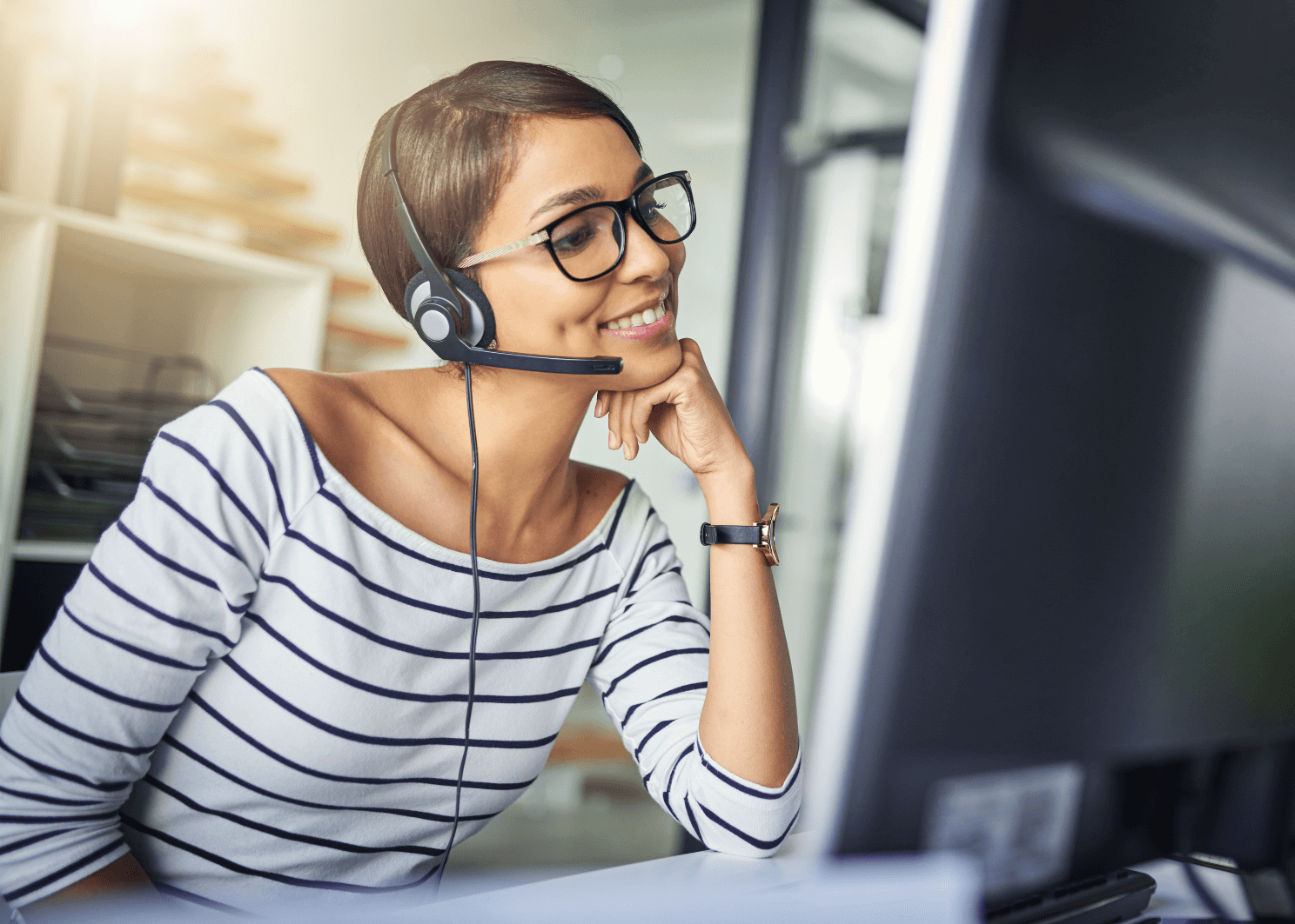 Smiling woman in a striped shirt wearing a headset sits at a desk in an office.