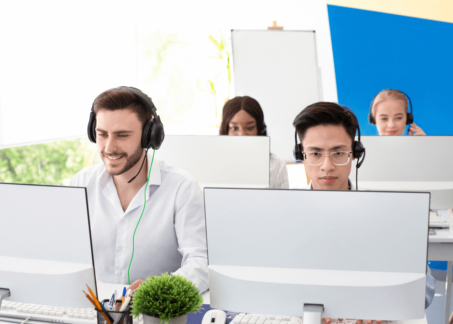 A team of customer service agents wearing headsets work at white desks with large computer monitors in a bright office.
