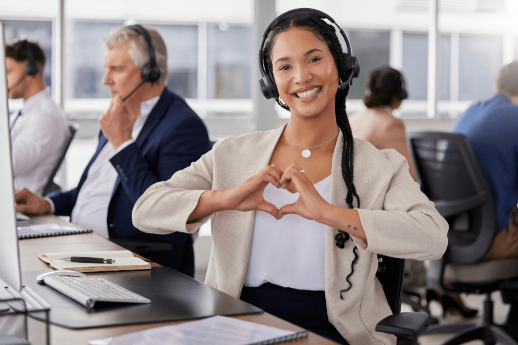 Smiling woman wearing a headset sits at a desk in a busy call center and forms a heart with her hands.