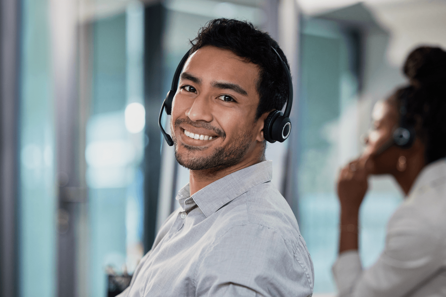 Smiling man wearing a headset in an office setting, with a coworker in the background.