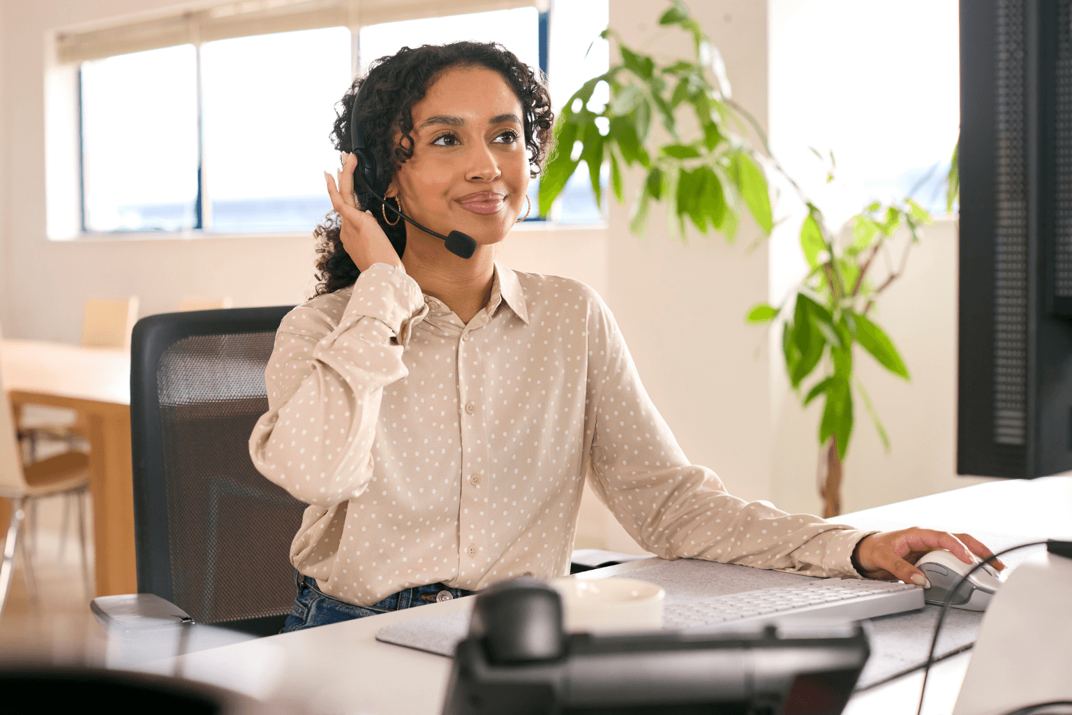 A woman wearing a headset sits at an office desk, talking on the phone with a plant in the background.
