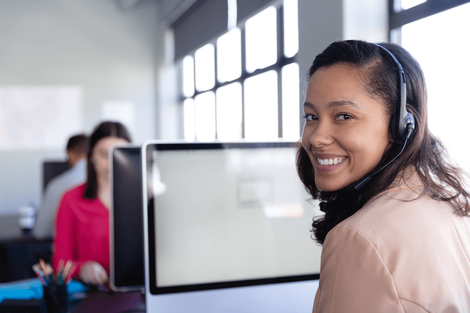 Smiling woman wearing a headset sits at a desk in a busy call center.
