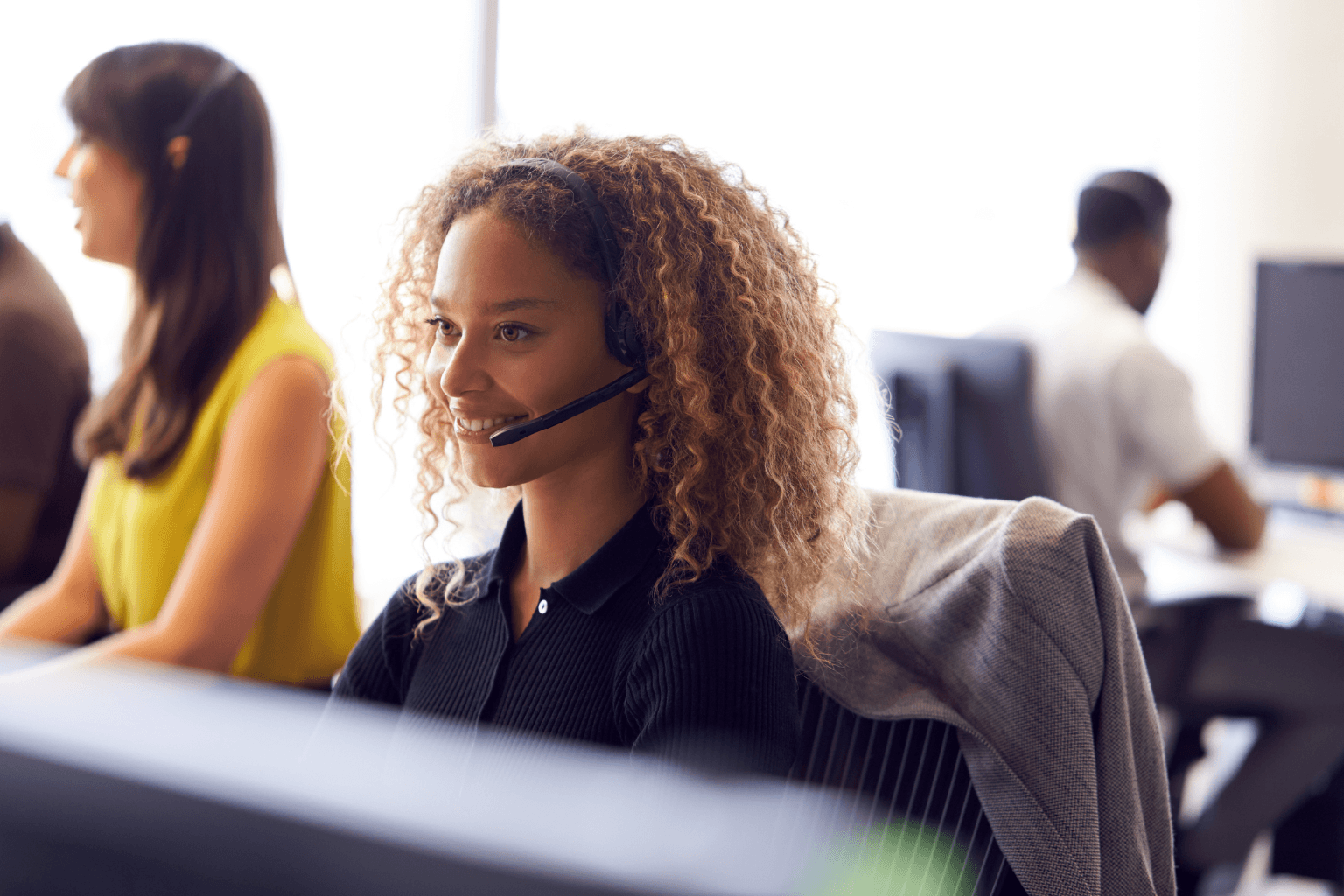 A woman with curly hair wearing a headset sits at a desk in a bright office, with coworkers in the background.