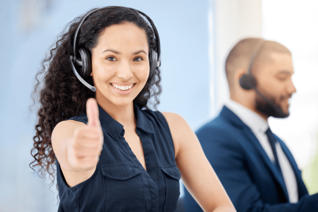 Smiling customer service representative wearing a headset gives a thumbs up, with a colleague in the background.