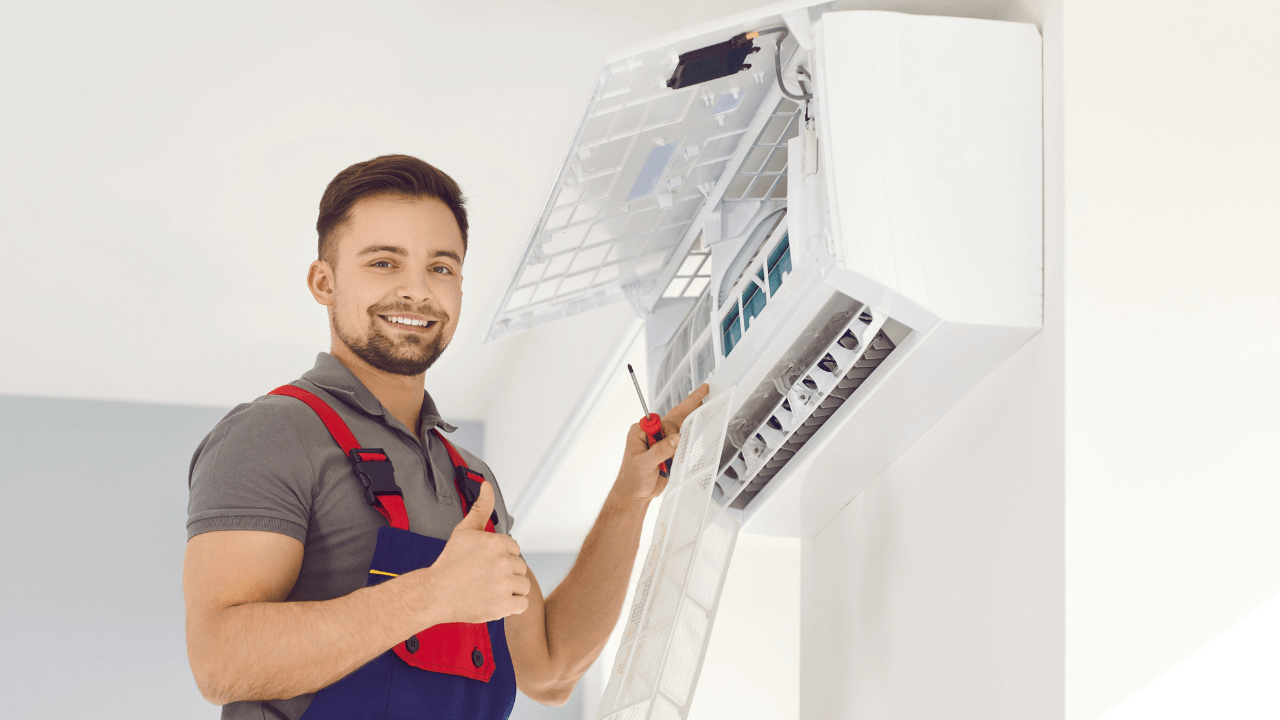 HVAC Technician in a gray shirt and red overalls giving a thumbs up beside an open air conditioner unit.