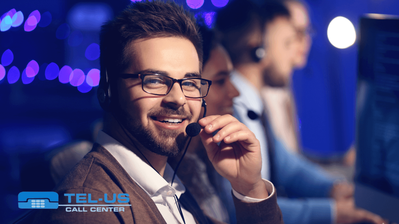 A smiling call center agent wearing a headset sits at a desk with computer monitors and other agents in the background.
