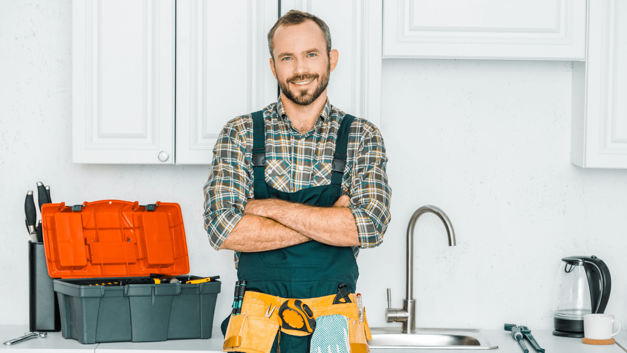 Smiling handyman in plaid shirt and overalls stands in a bright kitchen with a toolbox and tools on the counter.