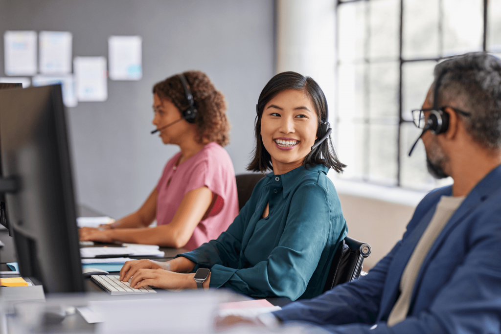 Smiling woman in teal sits at a desk with two colleagues using computers in a bright office.