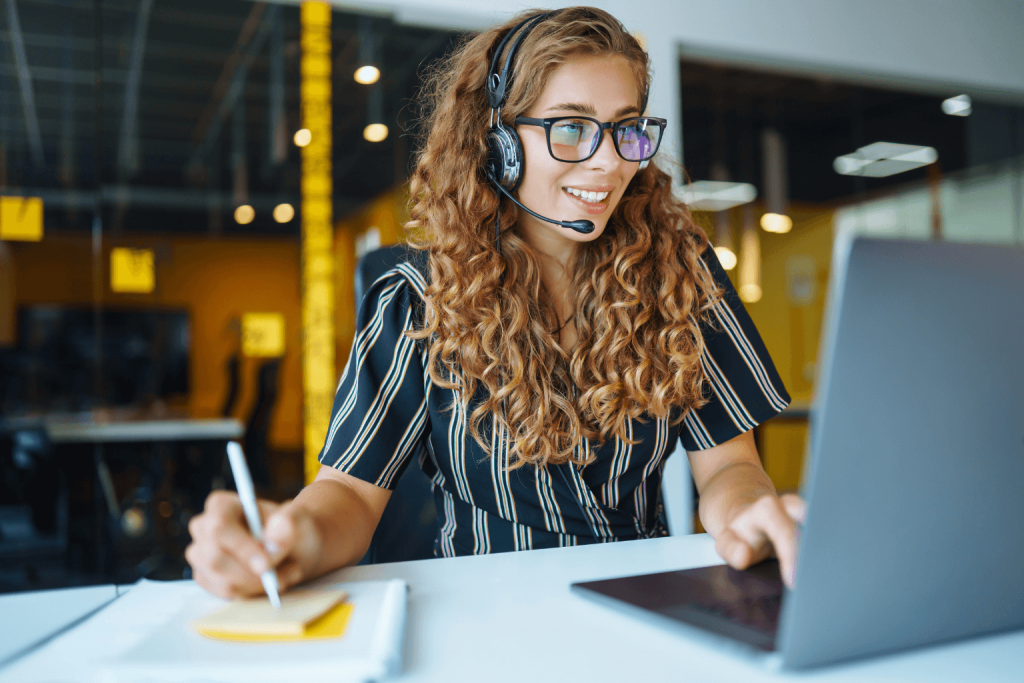 A smiling woman with curly hair wearing a headset sits at a desk with a laptop, taking notes on a yellow notepad in a bright office.