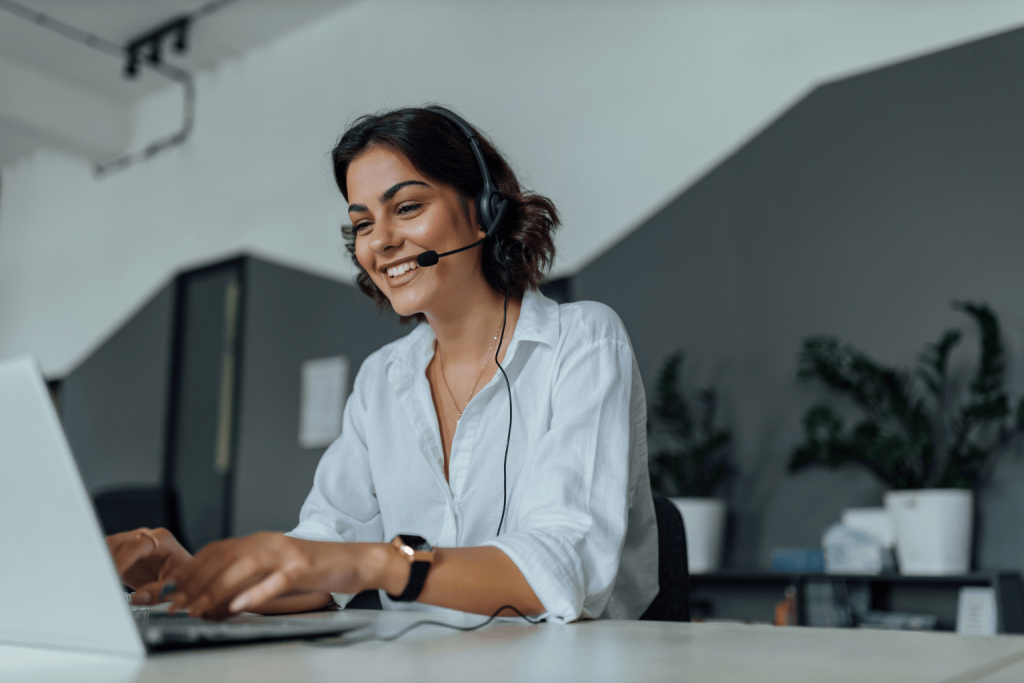 Smiling woman with a headset working on a laptop at a desk in an office.