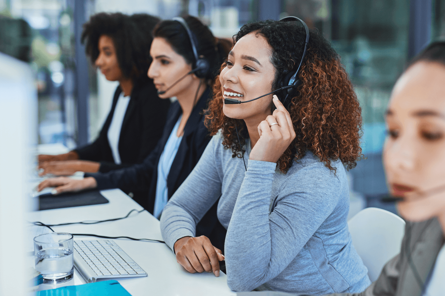 A diverse group of customer service representatives wearing headsets sit at desks with computers.