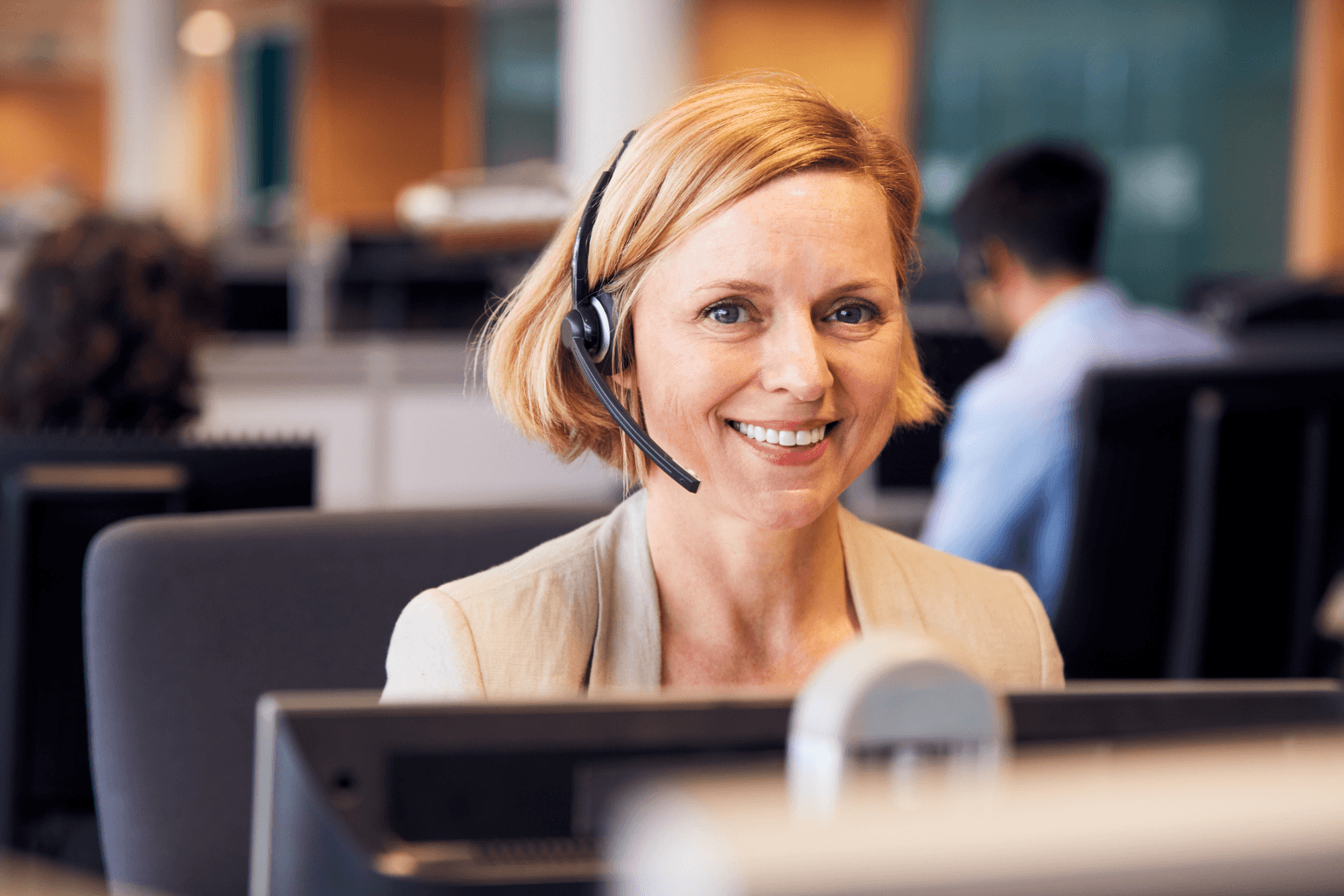 A smiling woman wearing a headset sits at a desk in a busy call center.