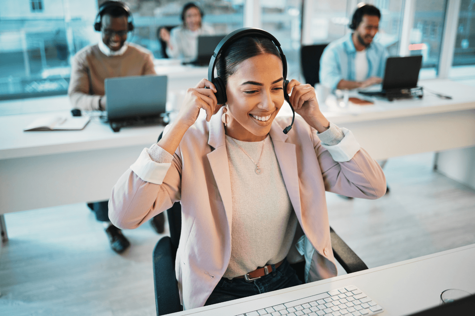 Smiling woman wearing a headset sits at a desk with a computer in a bright call center, with coworkers at their desks in the background.