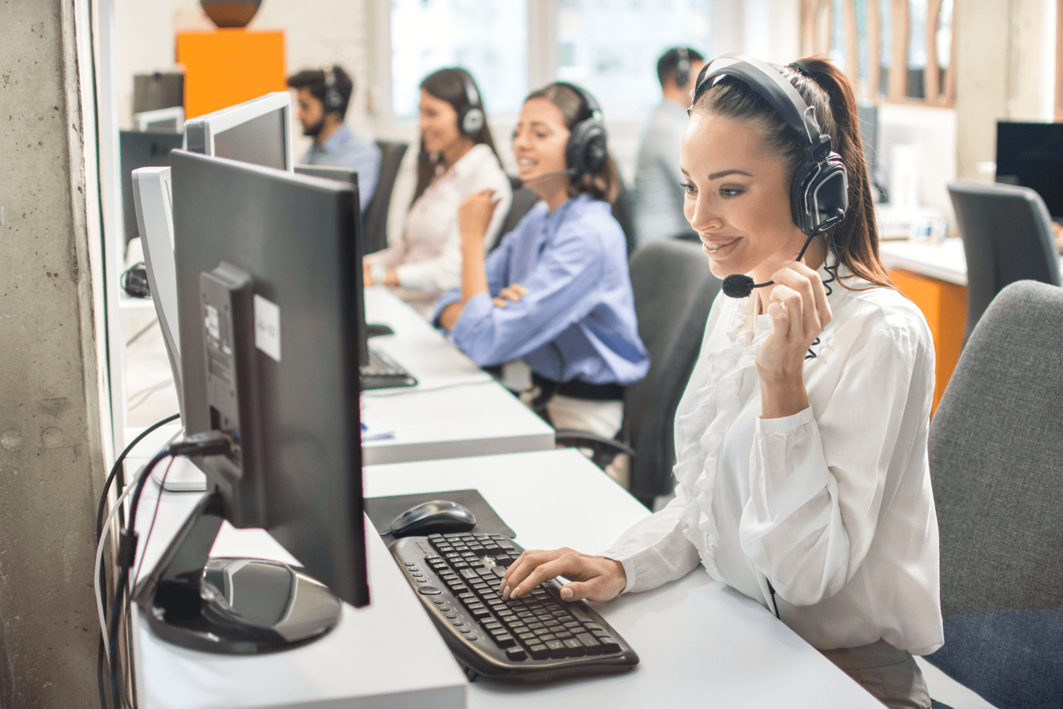 A woman wearing a headset works at a computer in an open-plan call center with coworkers at desks in the background.