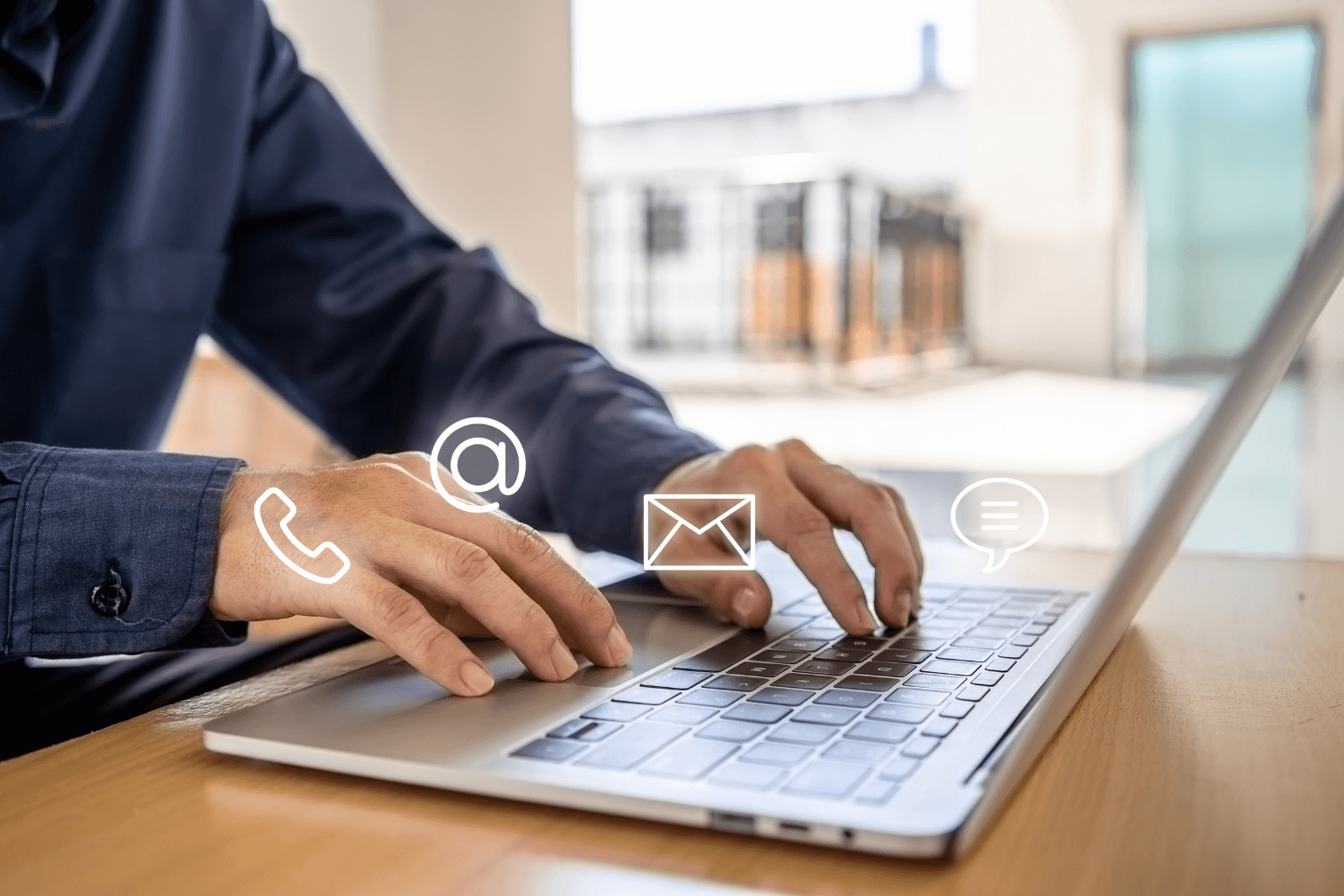 Person typing on a laptop at a wooden desk with floating communication icons for phone, email, @, and chat.
