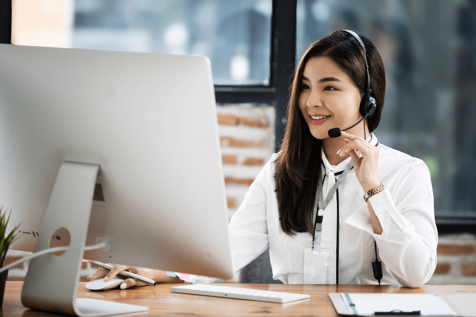 Woman wearing a headset sits at a desk with a large computer monitor, smiling in an office setting.