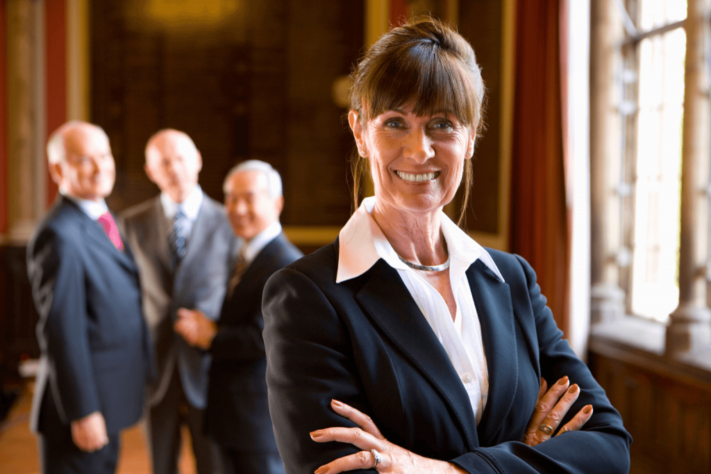 A confident lawyer in a dark blazer stands with arms crossed in the foreground as three suited colleagues chat in the background.