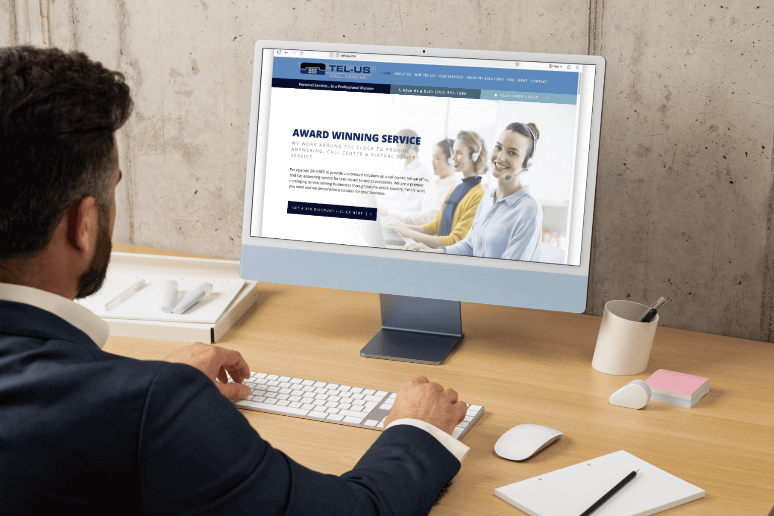 A man in a suit sits at a wooden desk, looking at a desktop monitor displaying a website with a smiling team photo.